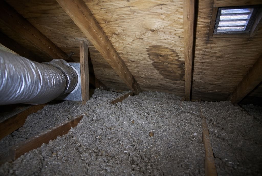 An older attic with thin loose-fill insulation, a bathroom fan duct and a water stain on the roof sheathing