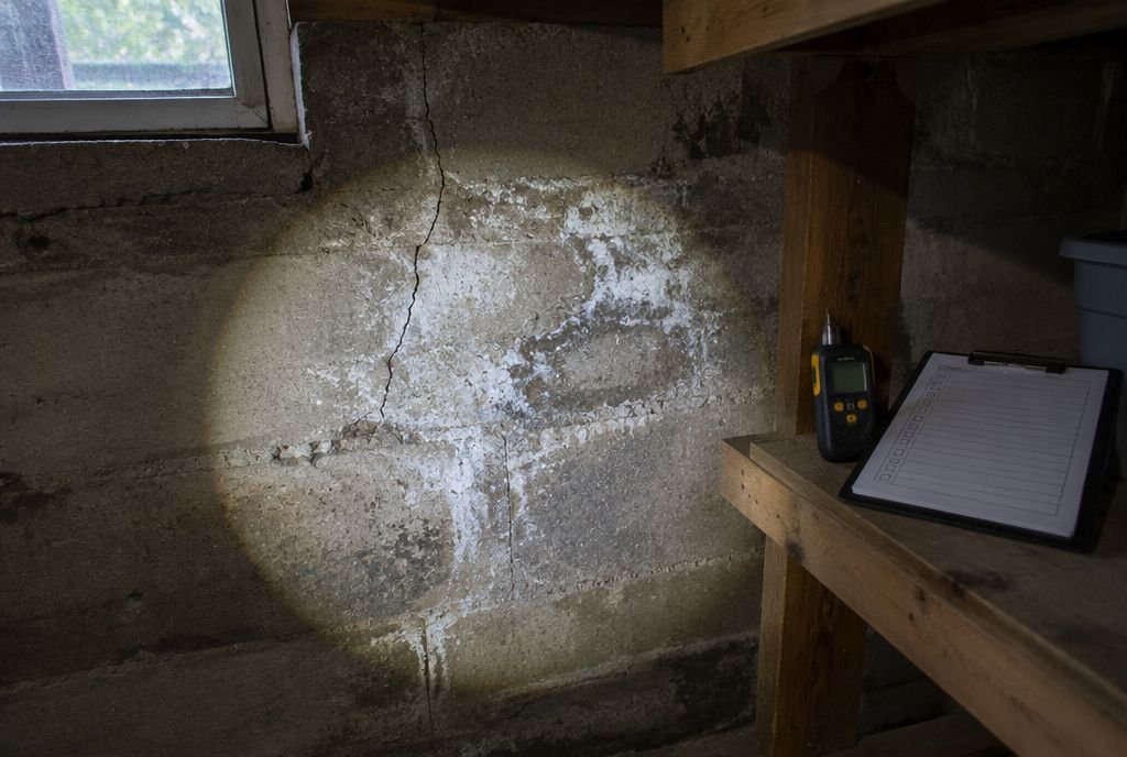 A flashlight on an older basement foundation wall showing a hairline crack and white efflorescence, with a moisture meter and clipboard nearby