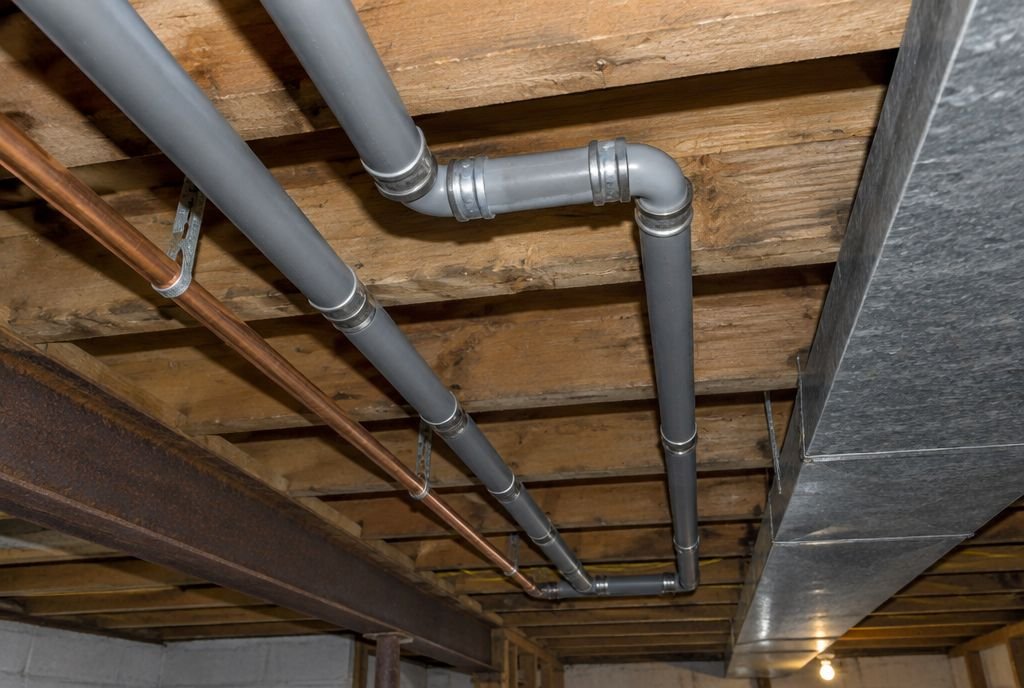 Grey polybutylene water pipes with crimped fittings running along basement floor joists beside copper and a duct